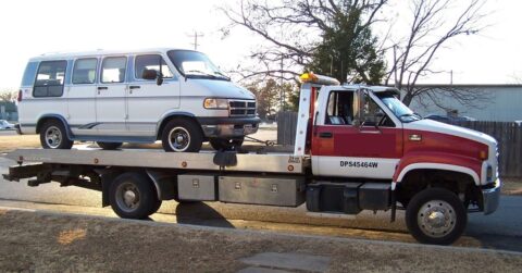 A red and white tow truck is transporting an old white passenger van, secured on its flatbed, on a suburban street.