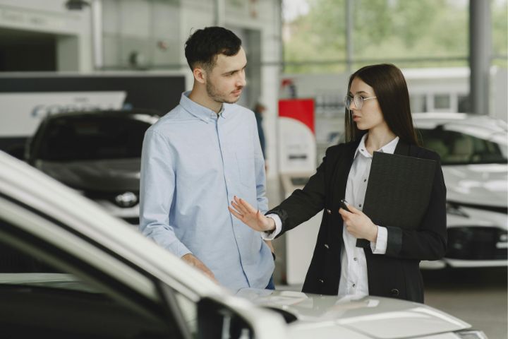 A car saleswoman gestures while explaining features of a white vehicle to a potential customer inside a bright, modern car dealership.