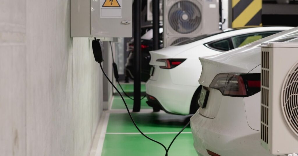 Two white electric cars are parked and charging in an underground parking garage, connected to wall-mounted charging stations near ventilation units.