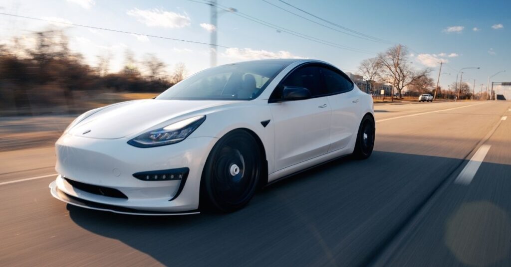A white Tesla Model 3 is seen driving at speed on a clear, open road with motion blur highlighting its movement.