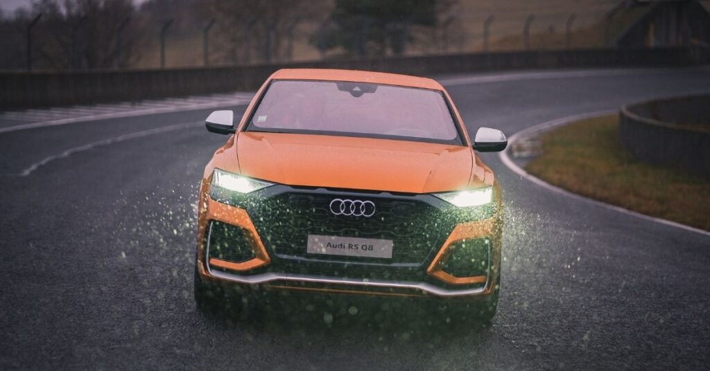 A yellow Audi RS Q8 with headlights on drives on a wet asphalt road, splashing water as it navigates a curve.