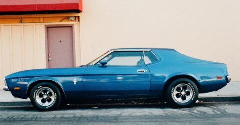 A vintage blue muscle car is parked on the street in front of a beige building with a pink door.