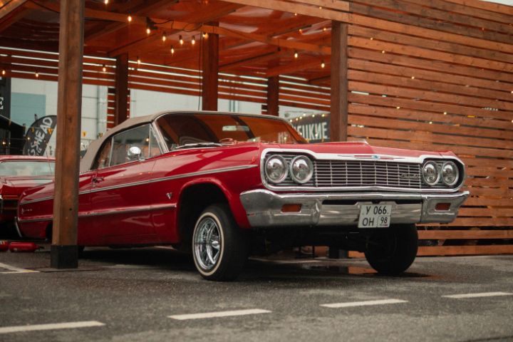 A classic red lowrider car with a raised front end is parked under a wooden canopy adorned with string lights.